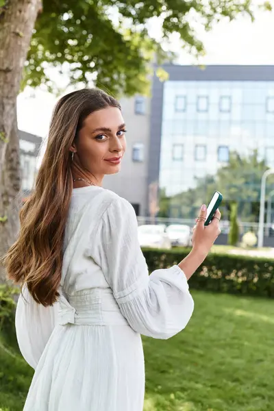 Young woman in a flowing dress stands outdoors with her smartphone, smiling in the sunlight. — Stock Photo