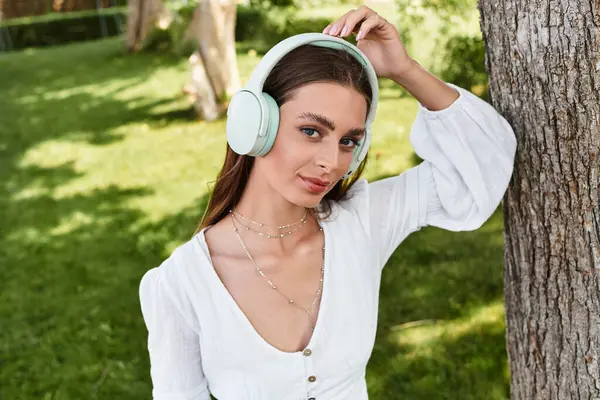 A beautiful young woman listens to music while relaxing by a tree in a vibrant park setting. — Stock Photo