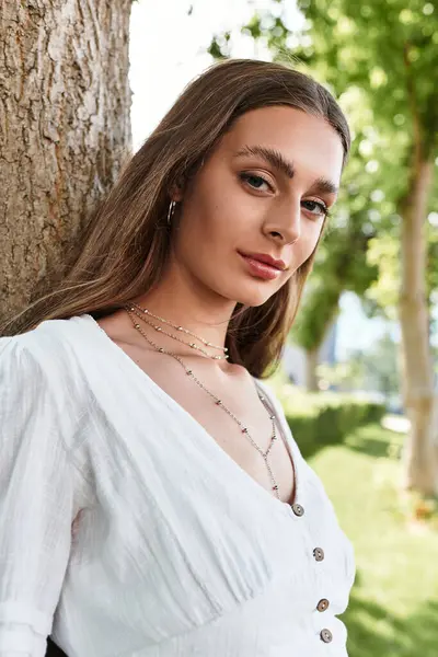 A beautiful young woman leans against a tree, showcasing her stylish outfit and radiant smile. — Stock Photo