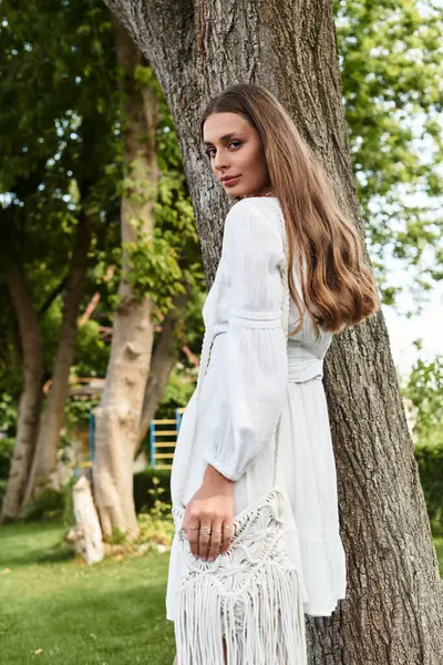 A young beautiful woman in a flowing white dress stands quietly beside a tree, exuding elegance. — Stock Photo