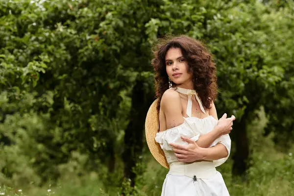 A beautiful young woman in a white dress stands gracefully in a lush park, enjoying nature's beauty. — Stock Photo