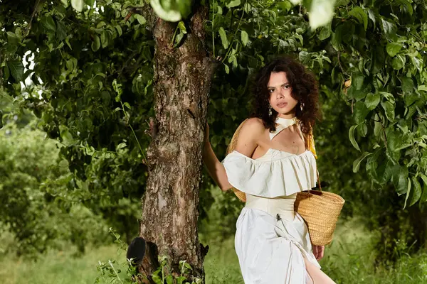 A young woman with curly hair stands gracefully against a tree while exploring a green park. — Stock Photo