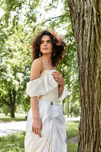A beautiful young woman in a white dress stands gracefully by a tree in a lush park. — Stock Photo