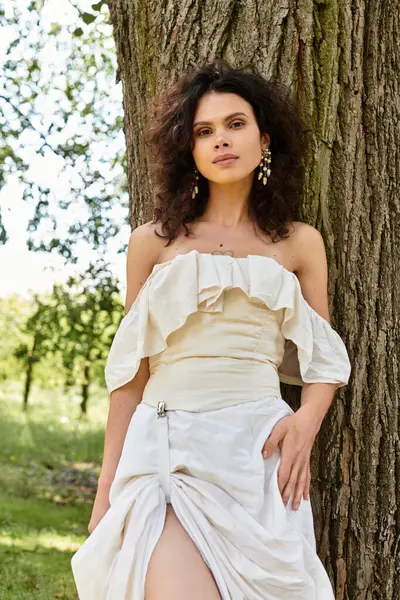 A young woman in a summer dress stands gracefully by a tree, surrounded by greenery. — Stock Photo