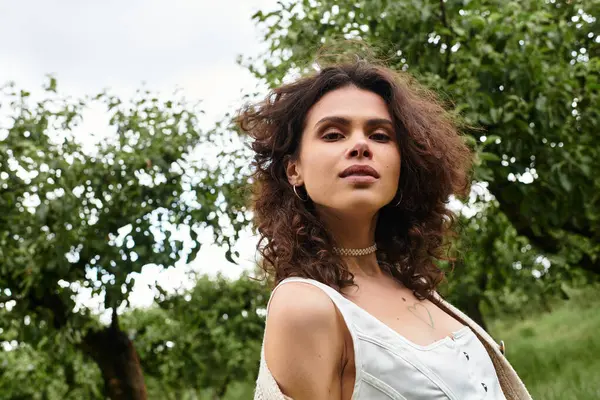 Curly-haired woman in a stylish summer outfit stands in a lush park surrounded by trees. — Stock Photo