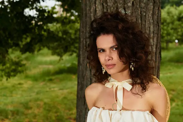 A young woman with curly hair stands gracefully in a lush green park wearing a lovely summer dress. — Stock Photo