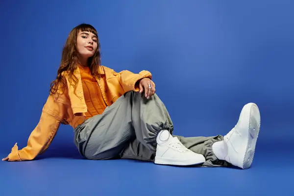 Une jeune femme à la mode s'assoit en toute confiance sur le sol portant des vêtements de rue élégants et des baskets. — Photo de stock