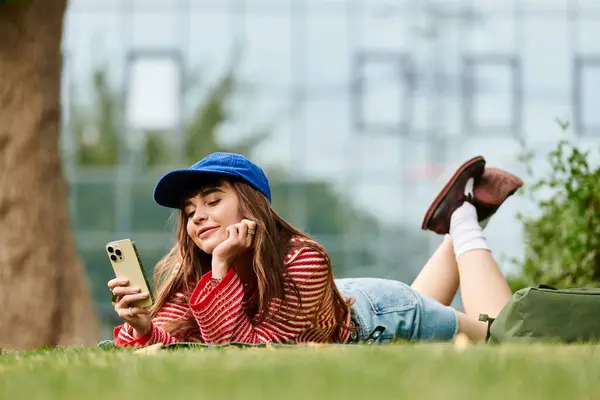 Веселая студентка расслабляется на траве, используя смартфон, пока учится на улице.. — стоковое фото