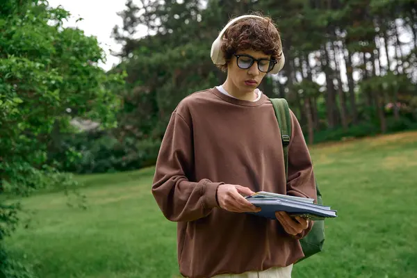 Молодой студент прогуливается по яркому зеленому парку, погрузившись в учебные материалы и музыку. — стоковое фото