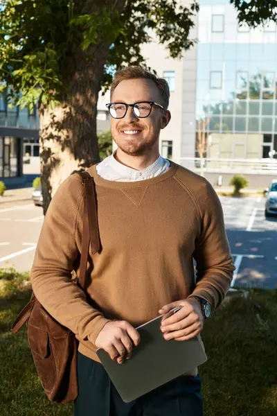 Young teacher stands outside a university building, smiling and holding a laptop under the sun. — Stock Photo
