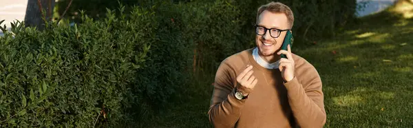 A cheerful young university teacher smiles while chatting on the phone in a vibrant garden. — Stock Photo