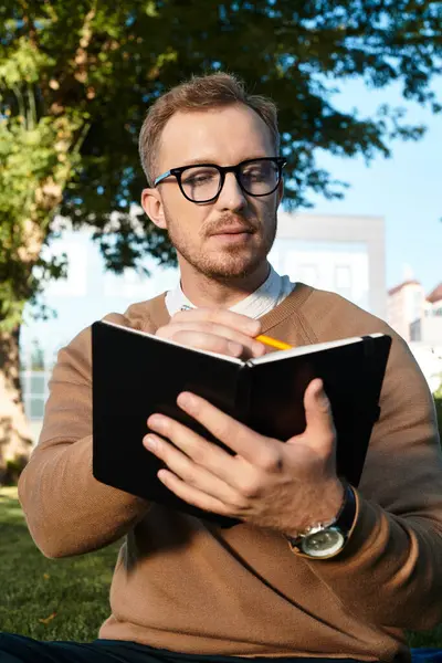 Un giovane insegnante si siede in un parco, scrivendo appunti in un quaderno, guardando premuroso. — Foto stock
