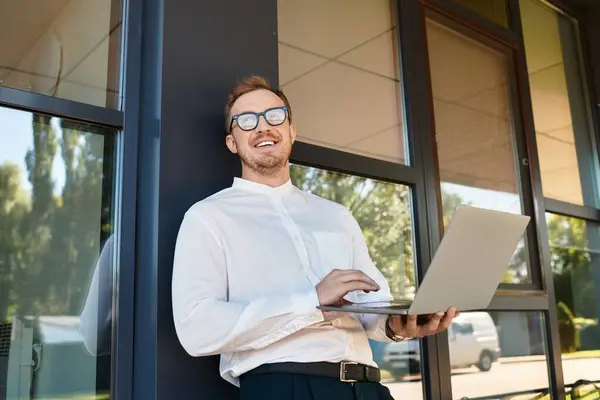 Молодой человек стоит у здания университета и улыбается, держа в руках ноутбук.. — стоковое фото