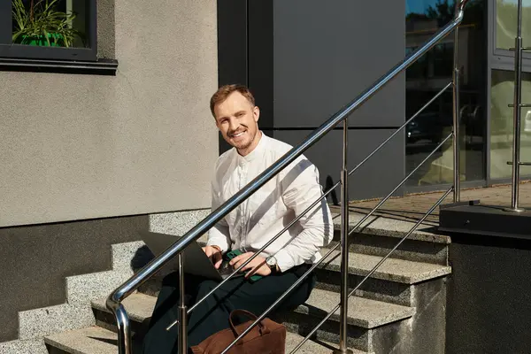 A young man dressed smartly sits on university steps, smiling while holding a laptop and a bag. — Stock Photo