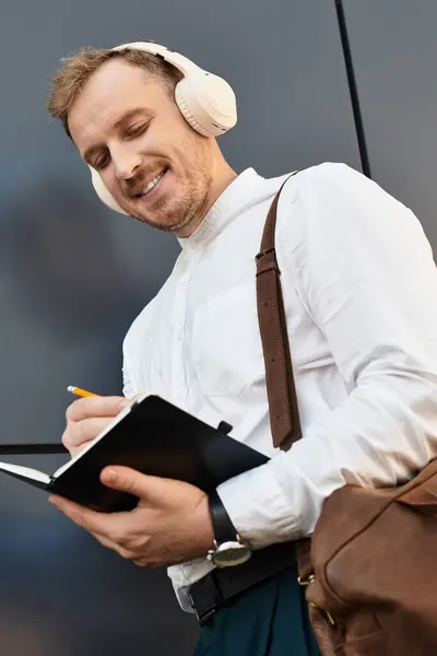 A young university teacher is writing notes in a notebook, wearing headphones and smiling. — Stock Photo