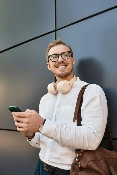 A young male teacher smiles at his smartphone, wearing headphones and carrying a bag. — Stock Photo