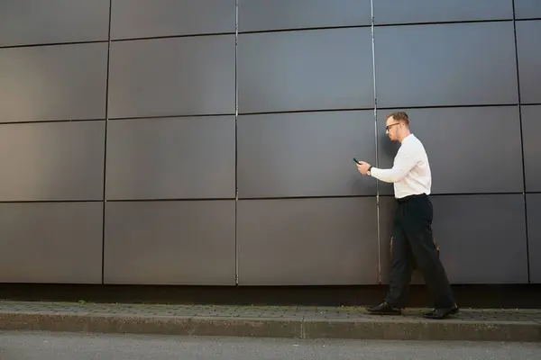 Молодой учитель в белой рубашке идет рядом с гладким университетским зданием, поглощенным телефоном. — стоковое фото