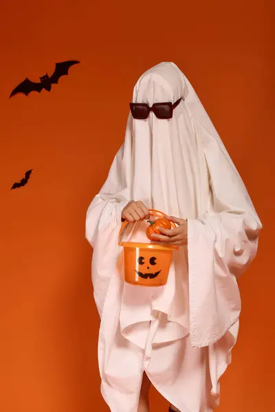 Uma criança vestida de fantasma celebra o Dia das Bruxas com um balde de abóbora contra um pano de fundo laranja. — Fotografia de Stock