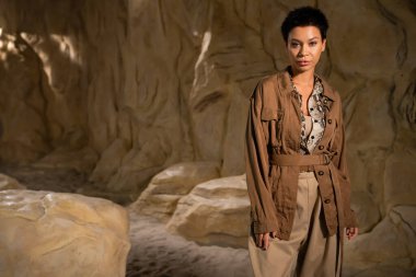 brunette archaeologist in brown jacket and beige pants looking at camera near rock in desert clipart