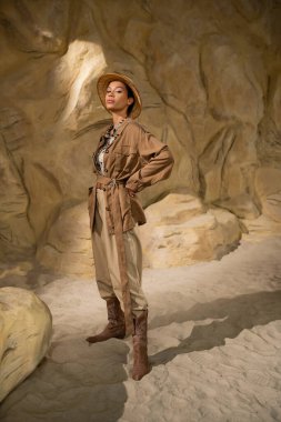 full length of archaeologist in safari hat and beige clothes standing with hand on hip in cave clipart