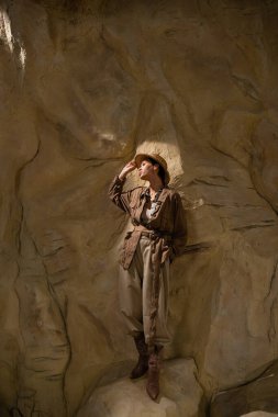 Safari tarzı giysiler içinde bir arkeolog çölde kayanın yanından uzağa bakıyor.