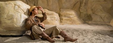 full length of young archaeologist sitting on sand near stone and adjusting safari hat, banner clipart