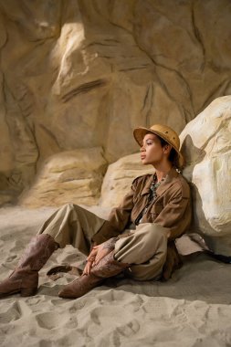 archaeologist in safari hat and beige clothes with cowboy boots sitting near stone in cave clipart