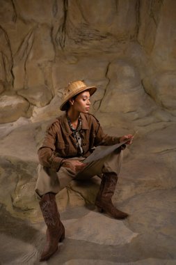 Safari şapkalı ve kovboy çizmeli arkeolog mağaradaki antik haritaya bakıyor.