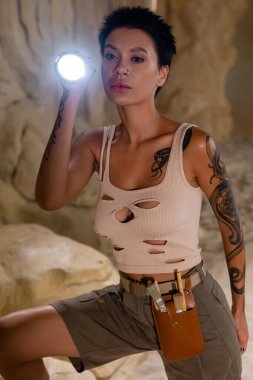Kolsuz tişörtlü seksi esmer arkeolog elinde el feneriyle çöl mağarasında duruyor.