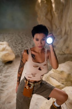 tattooed brunette archaeologist with waist bag holding flashlight in cave clipart