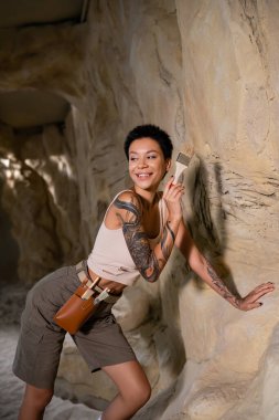 smiling tattooed archaeologist in tank top looking away while working with brush near rock in cave clipart
