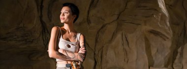 tattooed archaeologist in sexy tank top standing with crossed arms and looking away in cave, banner clipart