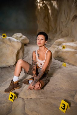 Tam boy seksi ve düşünceli arkeolog mağarada numaralandırılmış işaretlerin yanında oturuyor.