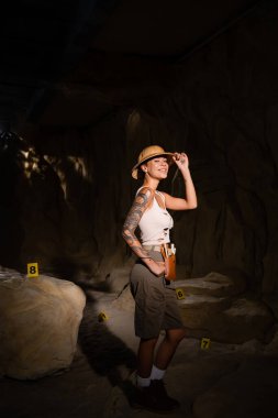 Seksi dövmeli arkeolog safari şapkasını ayarlıyor ve karanlık mağaradaki kartların yanındaki kameraya gülümsüyor.