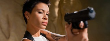 Esmer arkeolog çölde bulanık önplana silah doğrultuyor.