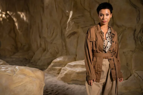 stock image brunette archaeologist in brown jacket and beige pants looking at camera near rock in desert