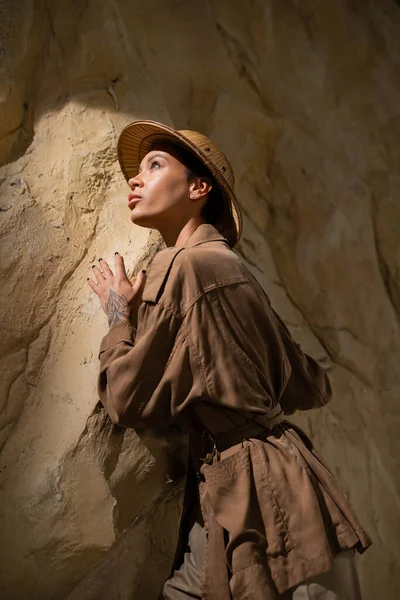 Bej ceketli ve safari şapkalı bir arkeolog mağaradaki kayanın yakınına bakıyor.