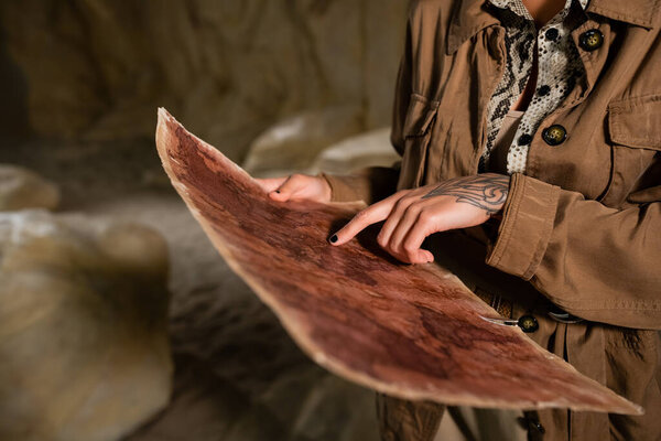 обрезанный вид археолога в коричневой куртке, указывающий на древнюю карту в пещере