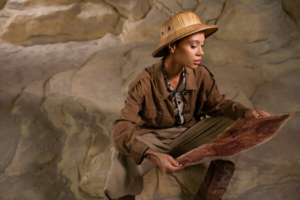 young archaeologist in safari style outfit sitting on rock and looking at ancient map