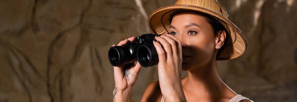 young and pretty archaeologist in safari hat holding binoculars and looking away in cave, banner