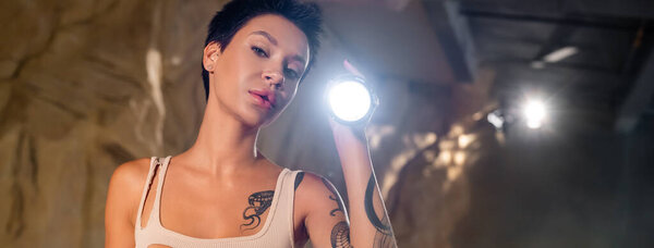 молодой татуированный археолог с фонариком, смотрящий в камеру в пещере, баннер