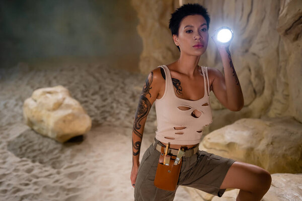 sexy archaeologist in tank top and waist bag with brushes holding flashlight in cave