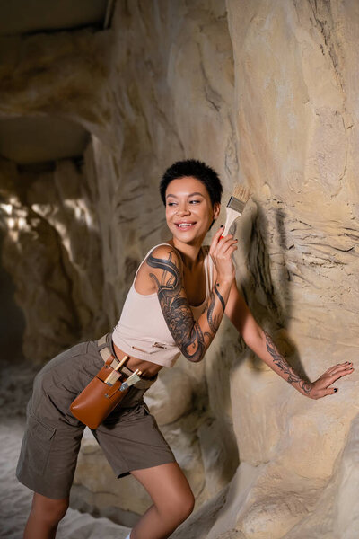 smiling tattooed archaeologist in tank top looking away while working with brush near rock in cave