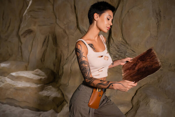 sexy archaeologist in tank top looking at ancient map near rock in desert