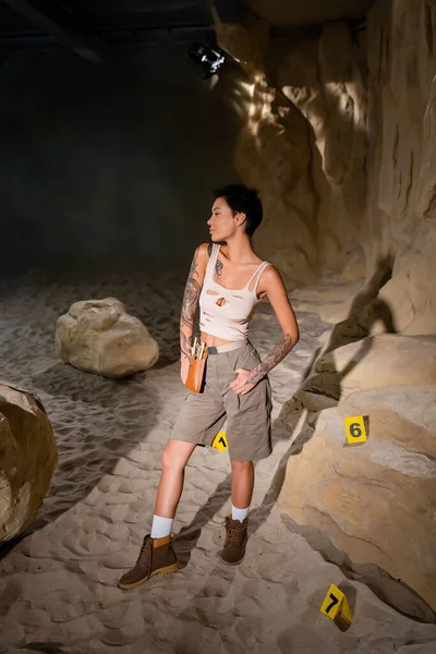 Uzun boylu, dövmeli bir arkeolog mağarada kumdaki kartların yanında duruyor.