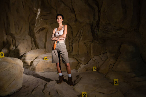 full length of young archaeologist standing with crossed arms near numbered marks in cave