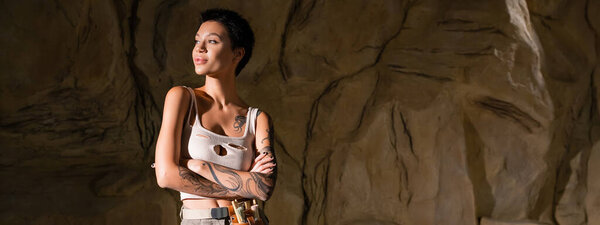 tattooed archaeologist in sexy tank top standing with crossed arms and looking away in cave, banner