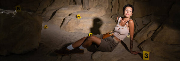 sexy archaeologist in tank top sitting near numbered cards in dark cave and looking away