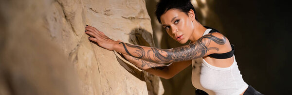 tattooed young archaeologist with shirt hair leaning on rock, banner