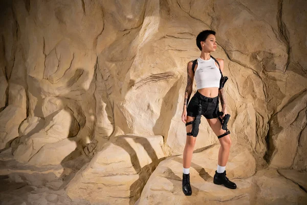 stock image full length of tattooed archaeologist in sexy outfit holding handgun and standing near rocks 